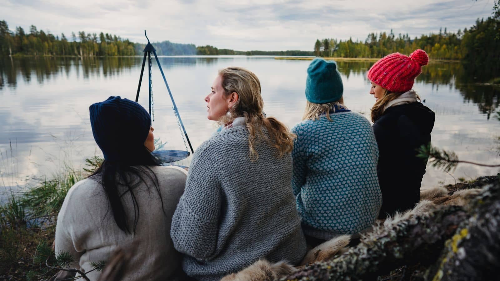 To personer sitter på en stein ved vannkanten med utsikt over en innsjø og skog i bakgrunnen. En kano med norske flagg ligger ved bryggen