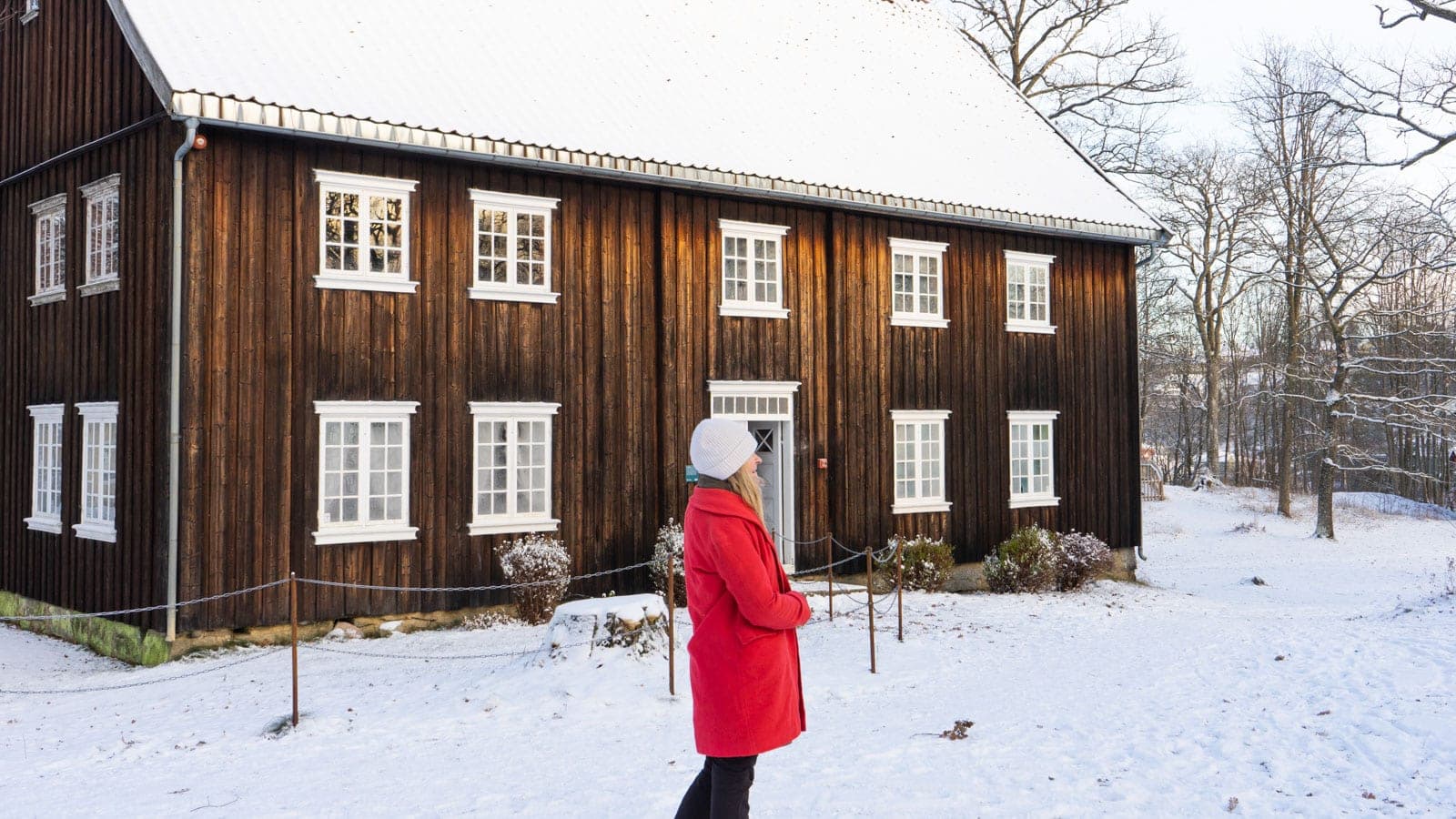 Gammelt trehus i vinterdrakt og en dame i rød kåpe som står utenfor på snøen