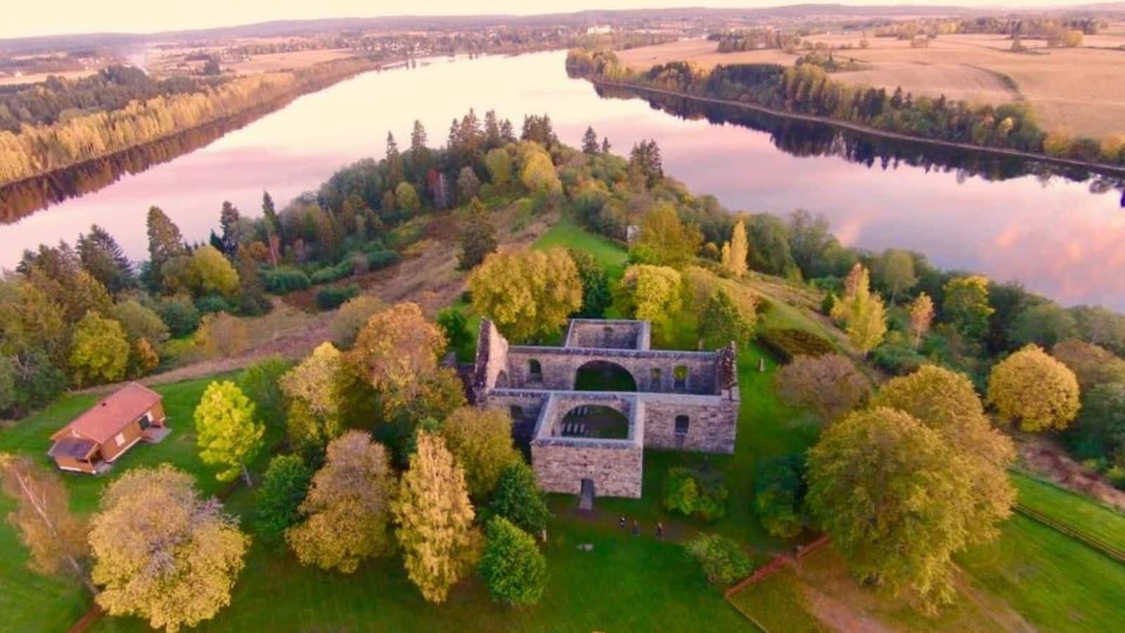 Dronebilde av Nes kirkeruiner omgitt av høstfarger, elvemøtet mellom Glomma og Vorma jorder og skog på Romerike