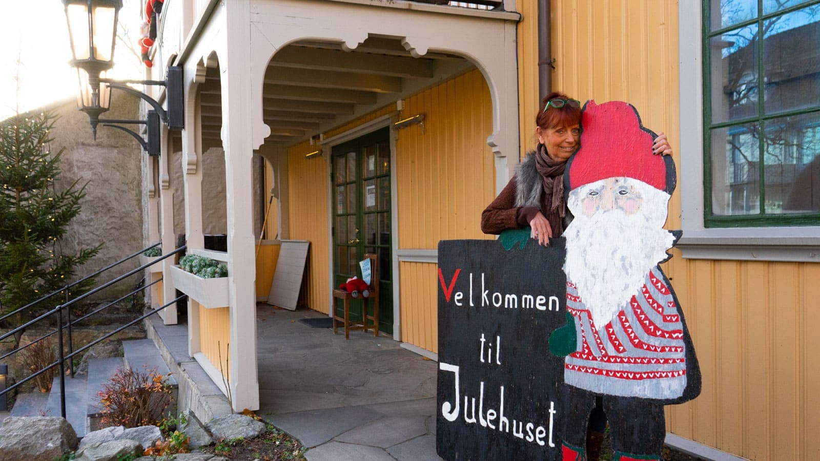 Dame som holder om et nisseskilt utenfor et gult trehus